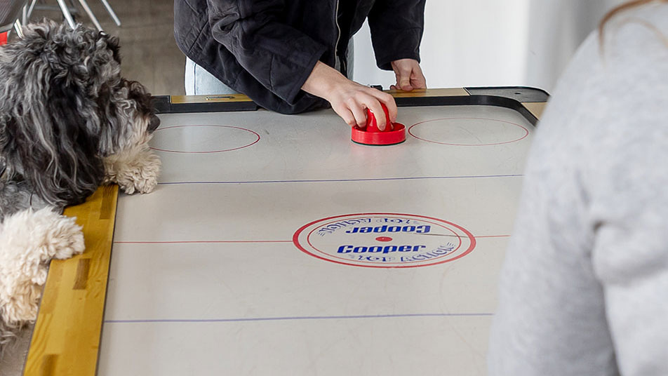 Two women play air hockey, with a dog named Toby perched on the edge of the table, involved in the action.