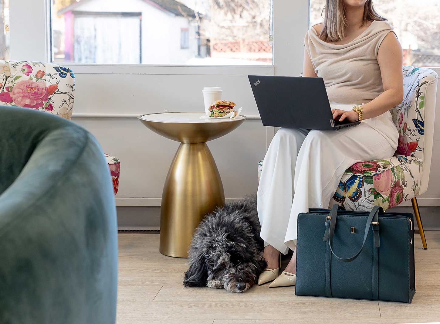 A wonderful grey and back dog sits nestled at the feet of a stylish business woman on a laptop. She is eating a sandwich, sitting on a floral print chair, with a blue briefcase at her feet. Her face is not visible.