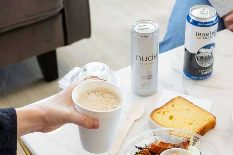 A person holds a paper cup of coffee in one hand, a bowl of fresh food with a slice of lemon cake for lunch. Nude vodka soda and a lager are visible in the background.