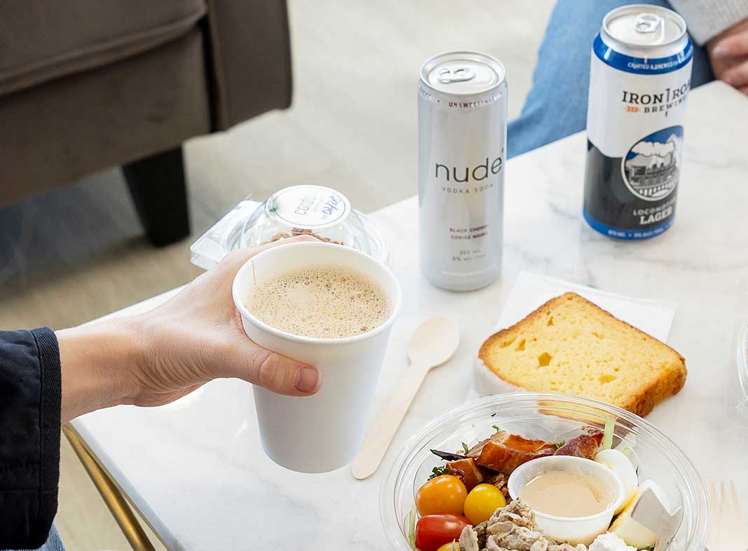 A person holds a paper cup of coffee in one hand, a bowl of fresh food with a slice of lemon cake for lunch. Nude vodka soda and a lager are visible in the background.