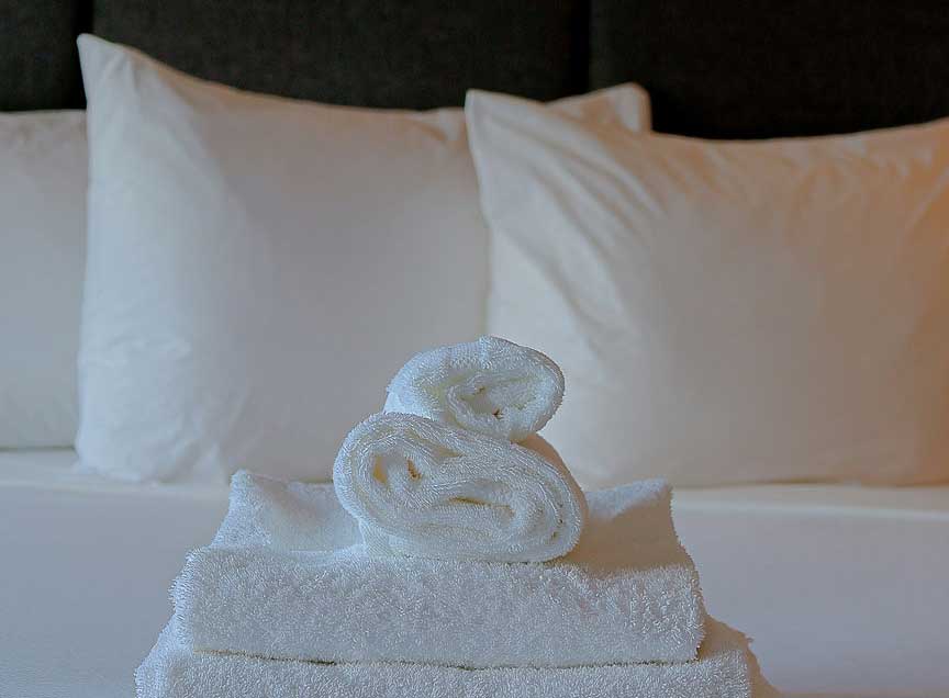 A rolled towel atop two others, on a white, clean hotel bed.