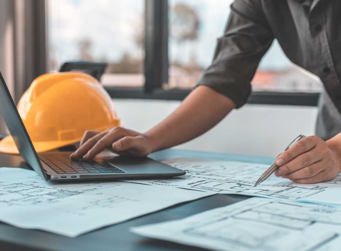 A government worker marks up a floor plan with one hand, edits on a computer with the other. A yellow hard hat is in the background.