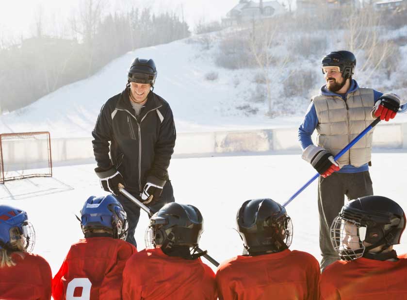 A team of hockey players are ready for the game, two coaches in front of them, outdoors on a sunny winter day.