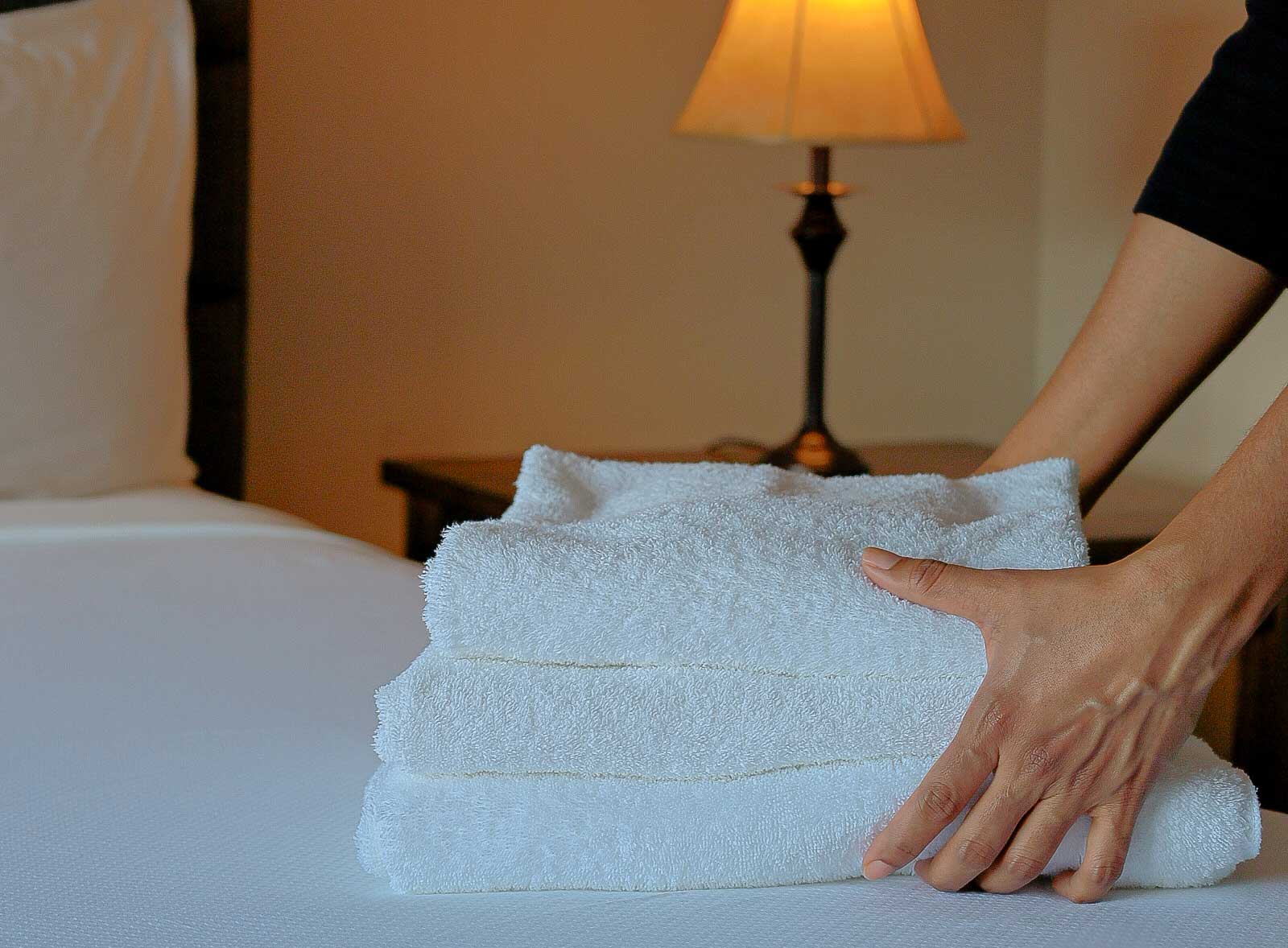 A woman's hands organize a pile of three white, clean towels on a hotel room bed.