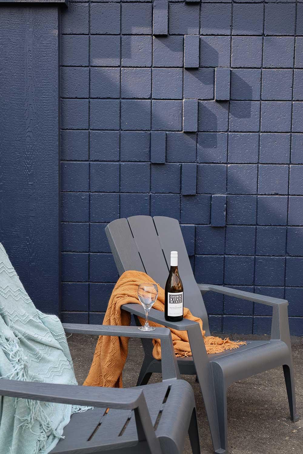 A grey chair, a wine glass, a wine bottle, and two blankets, one light blue and one orange. The wine bottle reads 