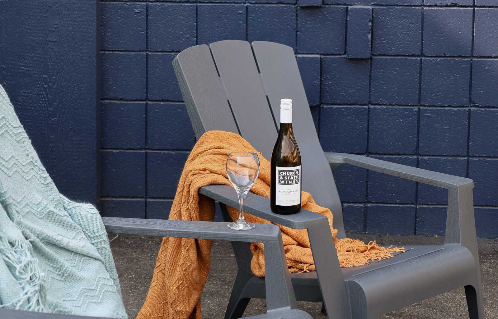 A grey chair, a wine glass, a wine bottle, and two blankets, one light blue and one orange. The wine bottle reads 