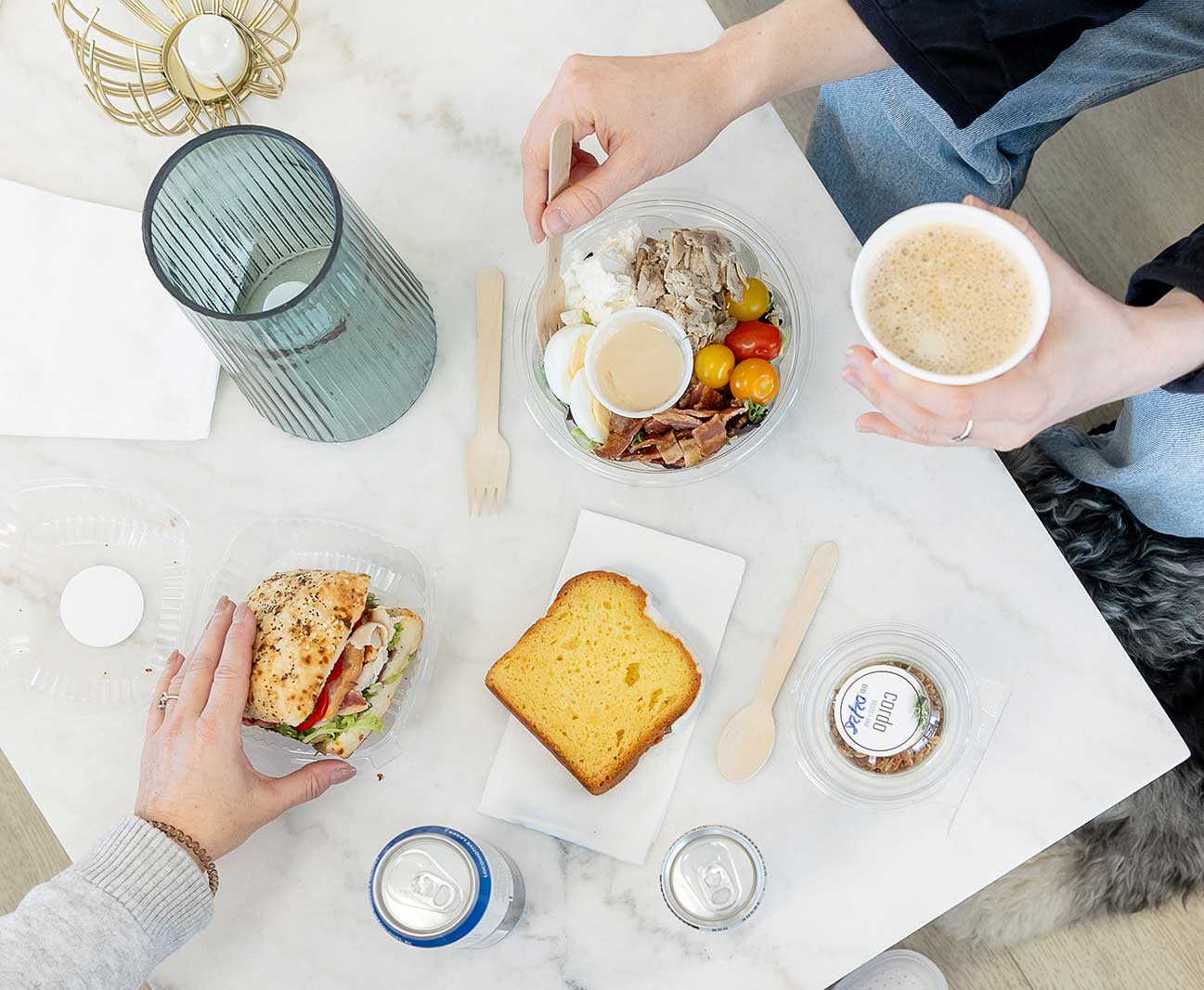 Two people enjoy lunch at Scott's Eats, complete with a bowl of fresh food, a sandwich, coffee, and a slice of lemon bread. The view is from above the table.